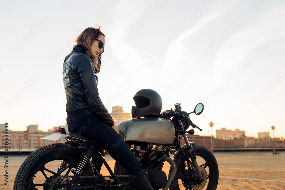 Sexy biker lady in black leather jacket sit on vintage custom caferacer  motorbike and touch her hair. Urban roof parking, sunset in big city.  Traveling and active hipster lifestyle. Girls power. –