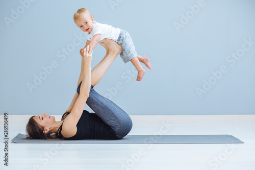 Sport, motherhood and active lifestyle concept - side view of mother workout together with her toddler boy over gray wall background. Mother having fun and playing with her little son. photo