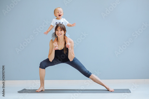 European sportive mother piggybacking her toddler baby son in fitness clothing on gray background. Lounge exercise. Motherhood, healthy lifestyle concept.