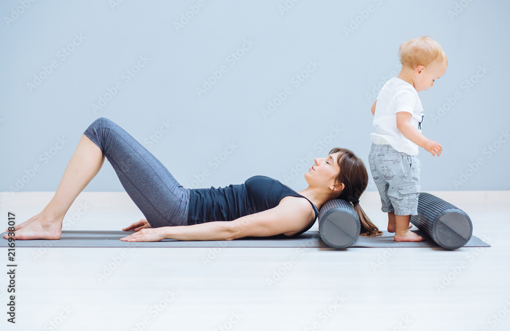 Obraz premium Young mother relaxing her neck and back with foam roller does physical pilates exercises while her toddler baby son playing near. Fitness, happy maternity sport with children concept.