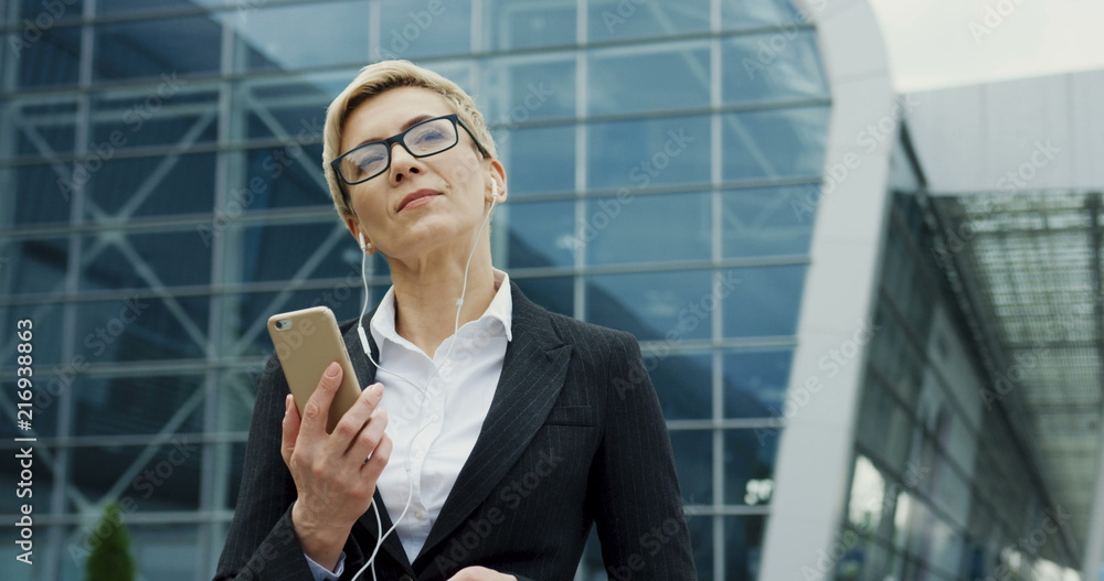 Caucasian middle aged woman with short hair, in glasses and headphones walking near big glass building and listening to the music on the smartphone. Caucasian middle aged woman with short hair, in glasses and headphones walking near big glass building and listening to the music on the smartphone.