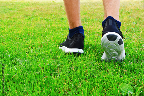 Man wearing black sneakers walking on green field in park and preparing to do workout and run closeup feet back side view with copy space for text on grass. Active lifestyle concept.
