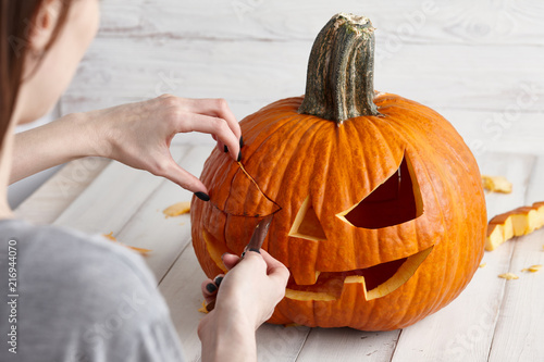 Woman carving big orange pumpkin into jack-o-lantern for Halloween holiday decoration on white wooden planks, close up view