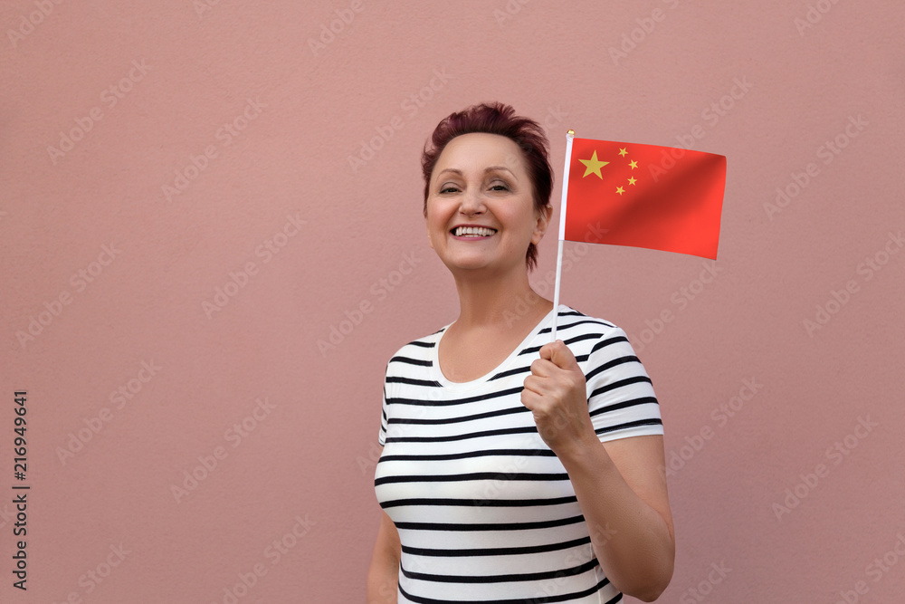 China flag. Woman holding Chinese flag. Nice portrait of middle aged pretty lady 40 50 years old with a national flag over pink wall background on the street. Learn Mandarin language concept. China flag. Woman holding Chinese flag. Nice portrait of middle aged pretty lady 40 50 years old with a national flag over pink wall background on the street. Learn Mandarin language concept.