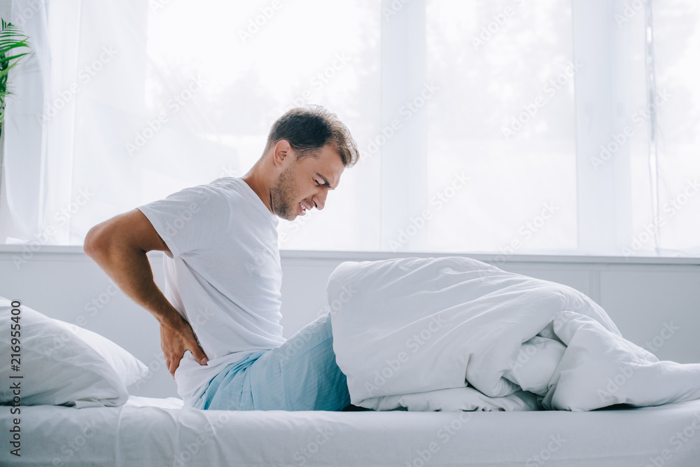 side view of young man in pajamas sitting on bed and suffering from back pain