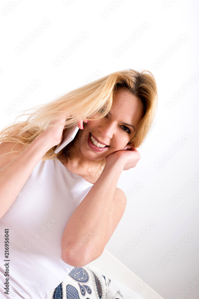 A beautiful smiling young woman sitting on a bed in a white tank top and talking on her phone. A beautiful smiling young woman sitting on a bed in a white tank top and talking on her phone.