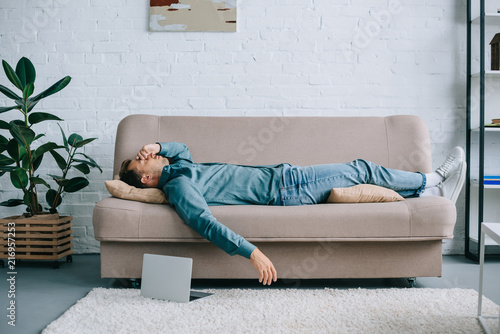 Photography young man with headache lying on sofa, laptop on carpet at home