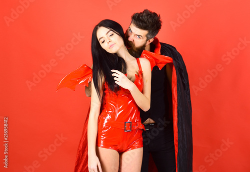 Couple in love play role game. Vampires victim concept. Man and woman dressed like vampire, demon, red background. Vampire in cloak behind sexy devil girl. Vampire bites female neck.