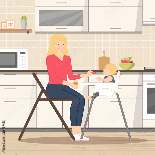 Young mother is sitting on the chair with little spoon and jar of baby puree in her hands, feeding her baby, who is sitting in a child's chair. Cozy kitchen interior background with furniture, stove.