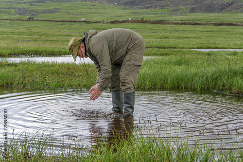 man in travel clothes scoops up water from a clean natural lake in the mountain tundra photo