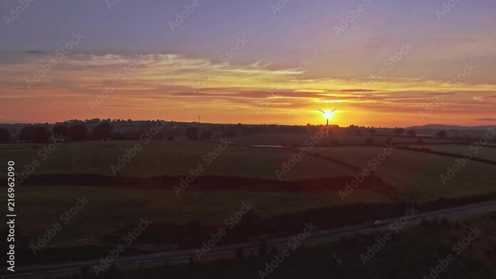 Car drives past as a silhouette of a windmill turns in background, just as the sun sets over the green hills of Ireland Car drives past as a silhouette of a windmill turns in background, just as the sun sets over the green hills of Ireland