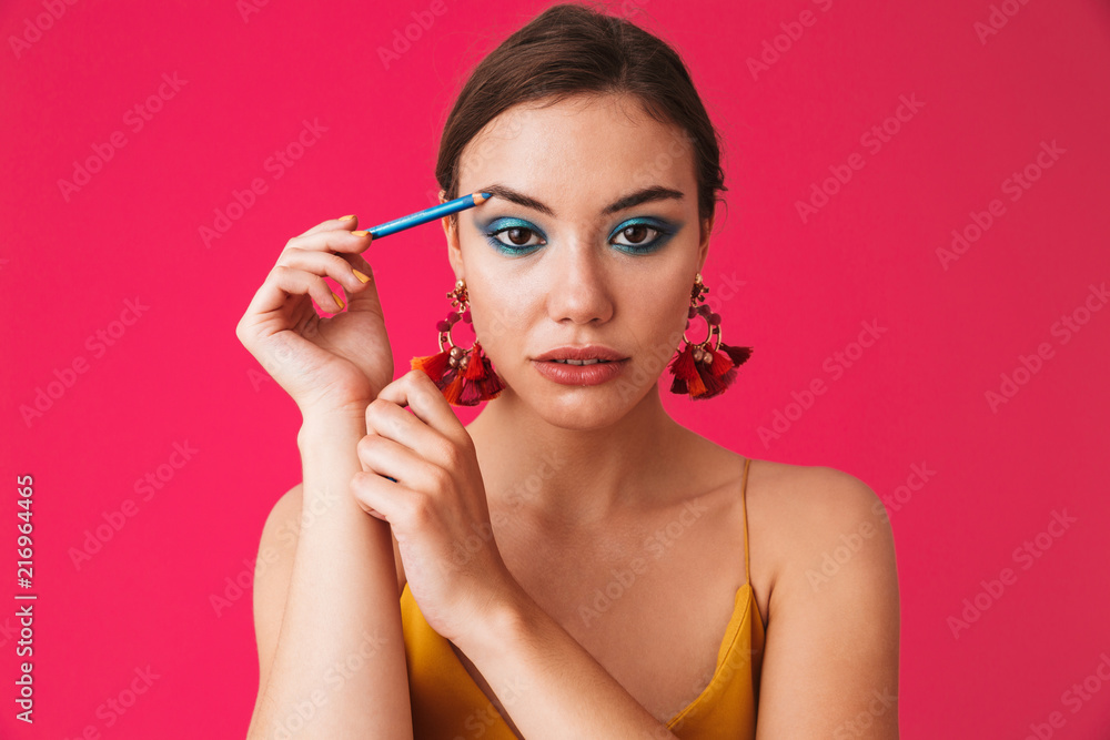 Fototapeta premium Image of caucasian stylish woman 20s wearing earrings smiling and applying makeup with blue eyeliner, isolated over pink background