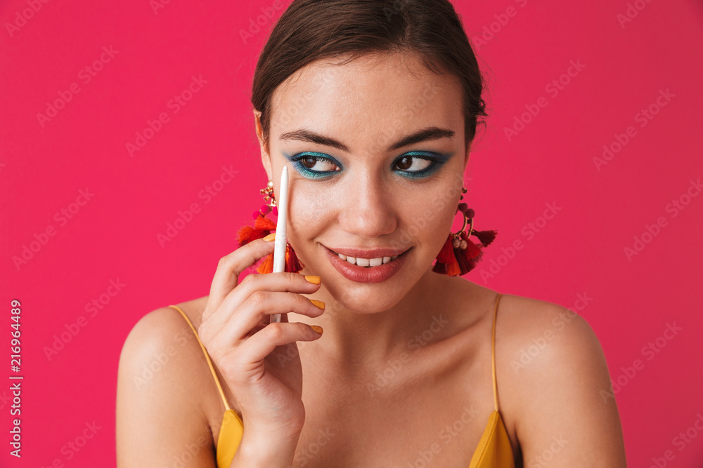 Photo of stylish trendy woman 20s wearing earrings looking aside and holding white pencil for makeup, isolated over pink background Photo of stylish trendy woman 20s wearing earrings looking aside and holding white pencil for makeup, isolated over pink background
