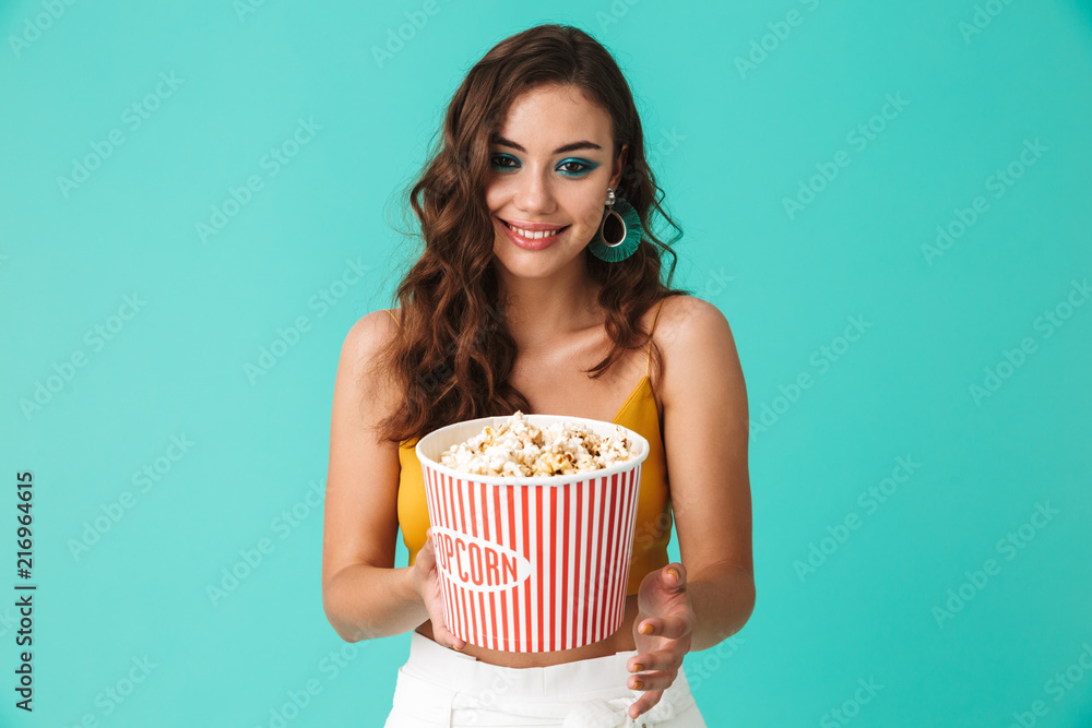 Photo of glamorous brunette woman 20s wearing fashion earrings holding bucket with popcorn and looking at camera, isolated over blue background