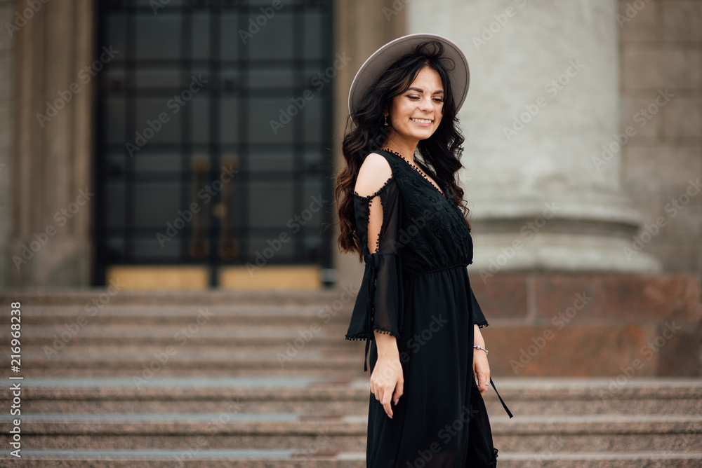 Naklejka premium Young beautiful stylish girl goes in a black dress in the city. Outdoor summer portrait of a young woman with a hat