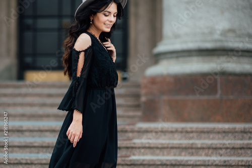 Young beautiful stylish girl goes in a black dress in the city. Outdoor summer portrait of a young woman with a hat