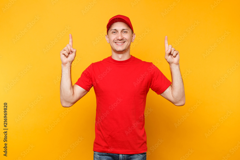 Delivery man in red uniform isolated on yellow orange background. Professional smiling male employee in cap, t-shirt working as courier dealer pointing index fingers up on copy space. Service concept.