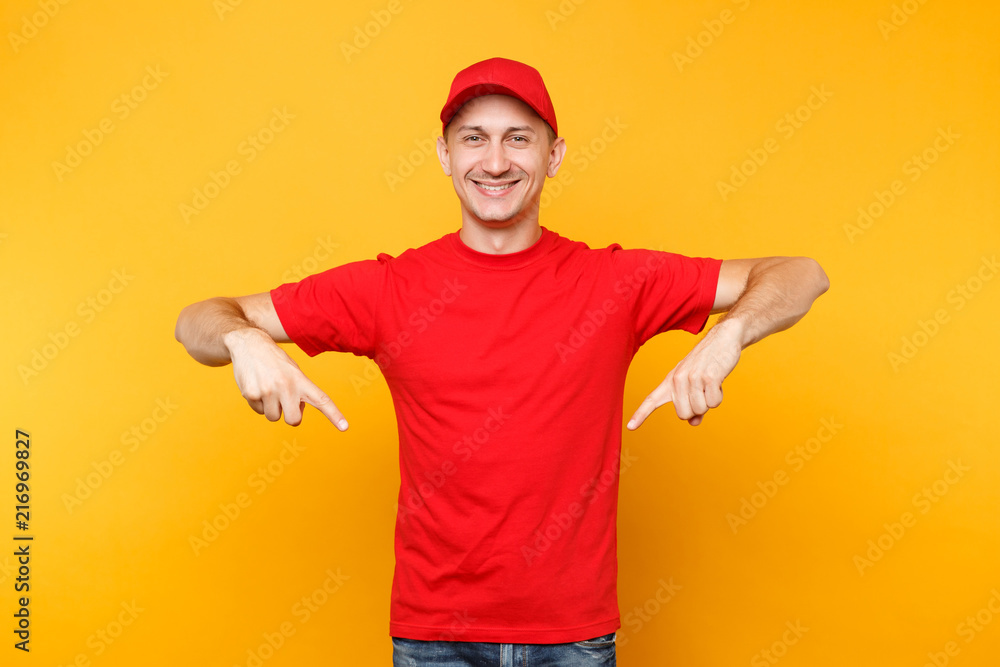 Delivery man in red uniform isolated on yellow orange background. Professional smiling male employee in cap t-shirt working as courier dealer pointing index fingers down on copy space. Service concept
