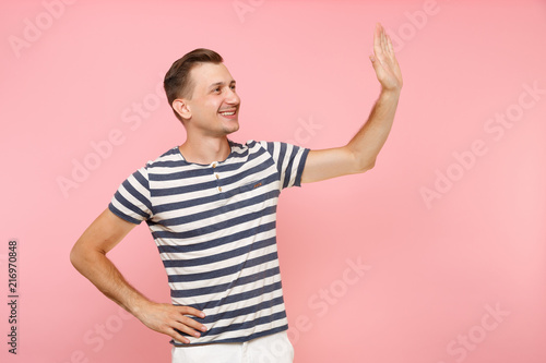 Portrait of smiling young man wearing striped t-shirt waving and greeting with hand as notices someone isolated on trending pastel pink background. People sincere emotions concept. Advertising area.