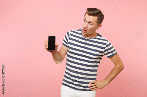 Portrait of man in striped t-shirt showing mobile phone camera with blank black empty screen copy space isolated on trending pastel pink background. People sincere emotions concept. Advertising area.