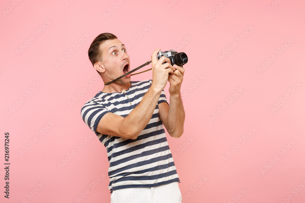 Portrait of surprised young photographer man wearing striped t-shirt take pictures on retro vintage photo camera isolated on trending pastel pink background. People sincere emotions lifestyle concept. - obrazy, fototapety, plakaty 