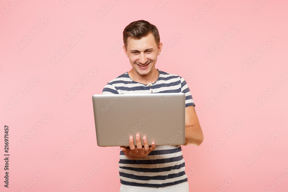 Portrait of handsome young man wearing striped t-shirt working on laptop computer, copy space isolated on trending pastel pink background. People sincere emotions lifestyle concept. Advertising area. Portrait of handsome young man wearing striped t-shirt working on laptop computer, copy space isolated on trending pastel pink background. People sincere emotions lifestyle concept. Advertising area.