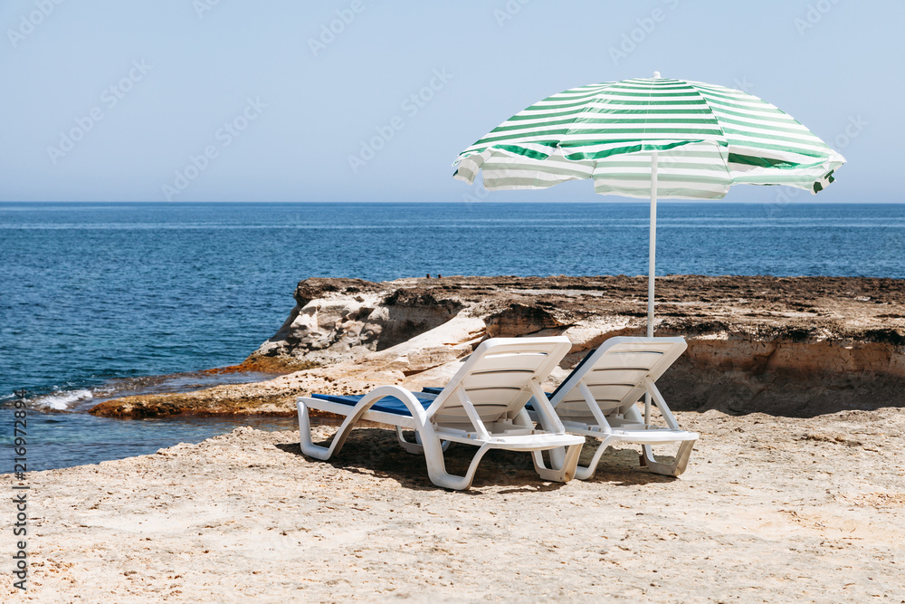Two blue deck chairs with a beach umbrella with green stripes in Malta / Two blue deck chairs with a beach umbrella with green stripes in a lonely beach.