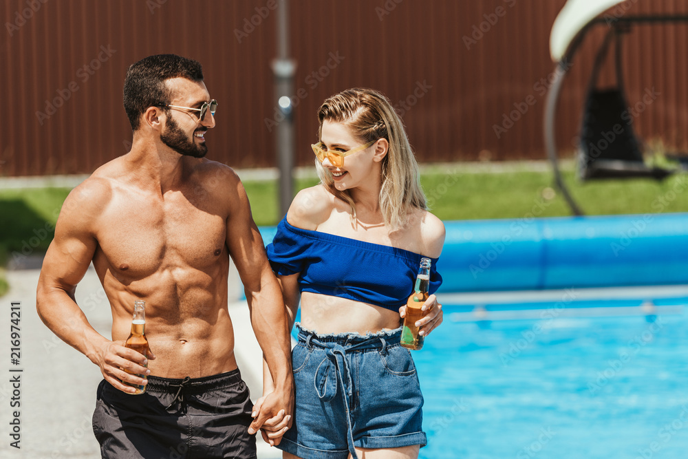 happy couple with bottles of beer holding hands at poolside happy couple with bottles of beer holding hands at poolside