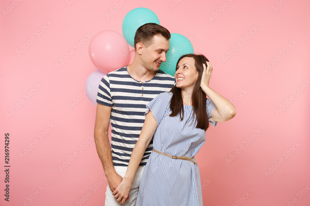 Portrait of young happy smiling couple in love. Woman and man in blue clothes celebrating birthday holiday party on pastel pink background with colorful air balloons. People sincere emotions concept.