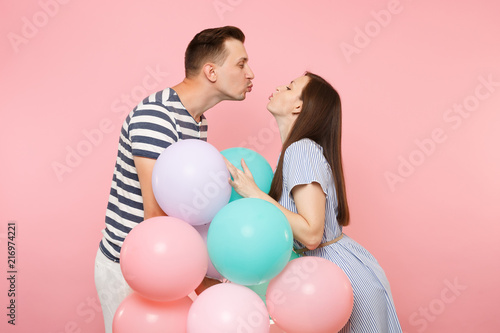Portrait of young happy kissing couple in love. Woman and man in blue clothes celebrating birthday holiday party on pastel pink background with colorful air balloons. People sincere emotions concept.