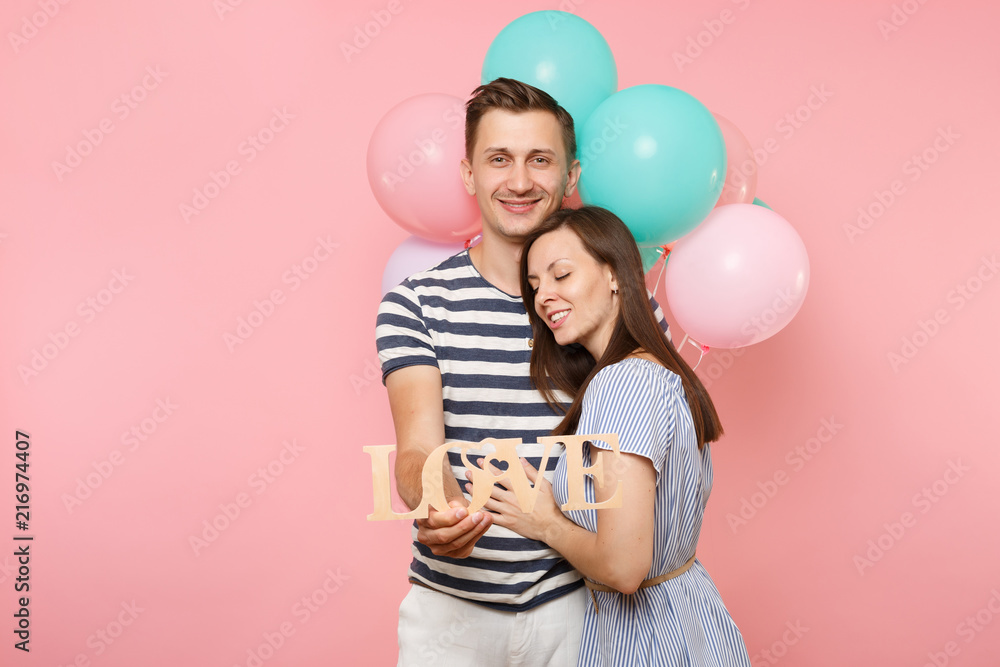Portrait of young happy smiling couple in love. Woman lean on one's man chest celebrating birthday holiday party on pastel pink background with colorful air balloons. People sincere emotions concept. Portrait of young happy smiling couple in love. Woman lean on one's man chest celebrating birthday holiday party on pastel pink background with colorful air balloons. People sincere emotions concept.