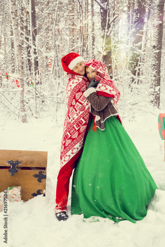 young couple, man and woman, husband and wife are walking in costumes of flowers typical of the elves of Santa's helpers in a winter forest under the snow with a chest full of gifts and a giant candy