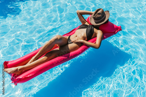 young woman in black swimsuit and straw hat sunbathing on pink inflatable mattress in swimming pool