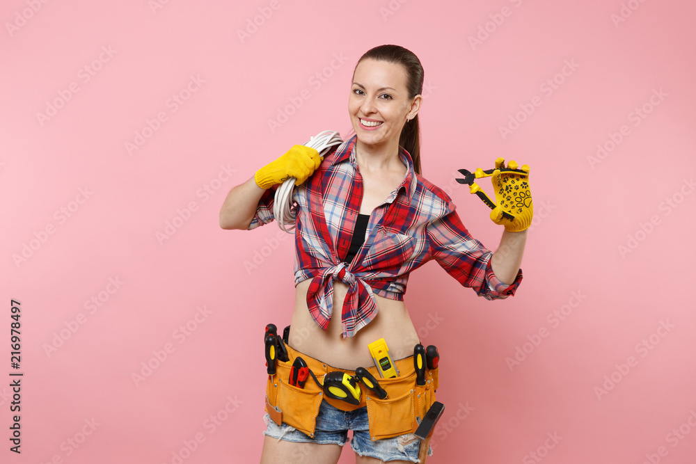 Young excited handyman woman in plaid shirt, denim shorts, kit tools belt full of variety instruments holding electric power cabel isolated on pink background. Female in male work. Renovation concept. Young excited handyman woman in plaid shirt, denim shorts, kit tools belt full of variety instruments holding electric power cabel isolated on pink background. Female in male work. Renovation concept.