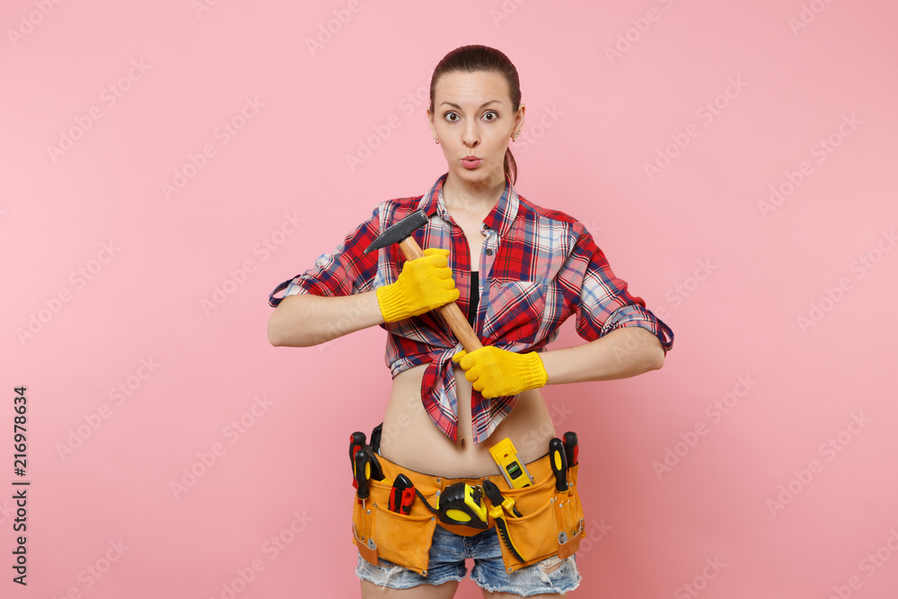 Strong young handyman woman wearing plaid shirt, denim shorts, kit tools belt full of different instruments hammer isolated on pink background. Female in male work. Renovation and occupation concept.