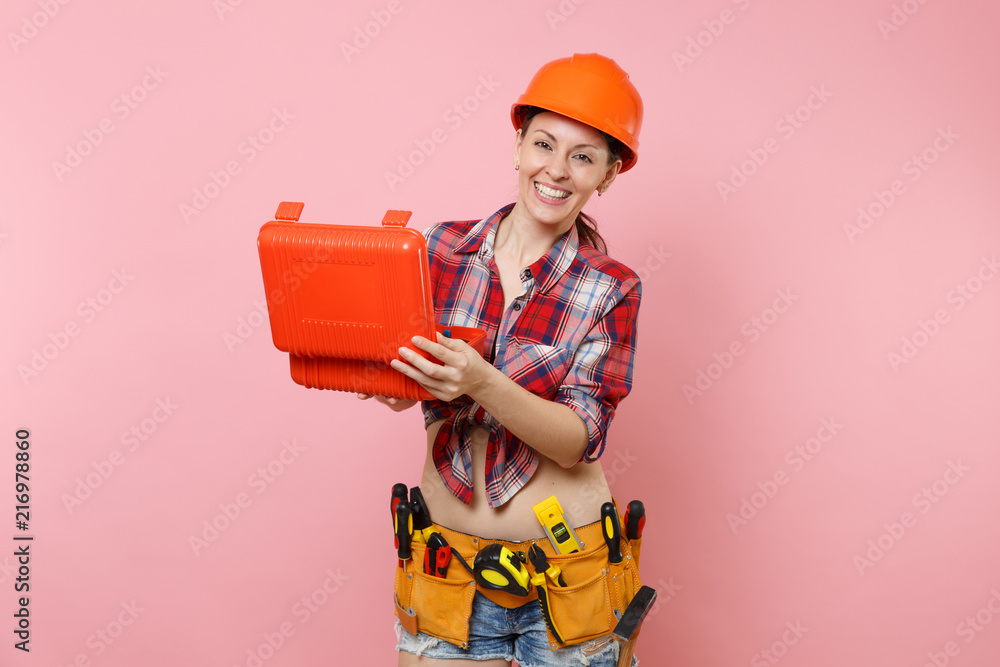 Strong young handyman woman in orange helmet, plaid shirt, denim shorts, kit tools belt full of instruments, toolbox isolated on pink background. Female in male work. Renovation and occupation concept Strong young handyman woman in orange helmet, plaid shirt, denim shorts, kit tools belt full of instruments, toolbox isolated on pink background. Female in male work. Renovation and occupation concept
