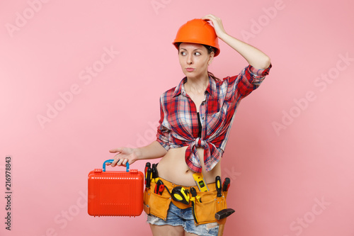 Strong young handyman woman in orange helmet, plaid shirt, denim shorts, kit tools belt full of instruments, toolbox isolated on pink background. Female in male work. Renovation and occupation concept © ViDi Studio