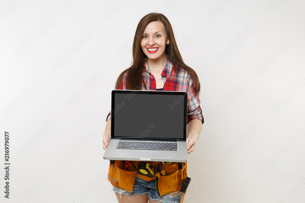 Woman in kit tools belt full of variety instruments holding laptop pc computer with blank black empty screen display monitor isolated on white background. Female doing male work. Renovation concept. Woman in kit tools belt full of variety instruments holding laptop pc computer with blank black empty screen display monitor isolated on white background. Female doing male work. Renovation concept.