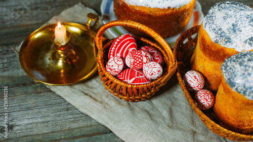 Red easter eggs with folk Ukrainian pattern lay into basket beside easter cakes and candlestick on sheet of vintage cloth on old wood background. Ukrainian traditional eggs pisanka and krashanka