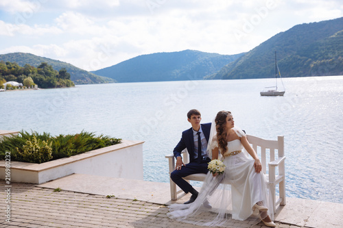 A stylish young bride and groom in a white lavish wedding dress are sitting on a bench in front of a river and hills. © romannoru