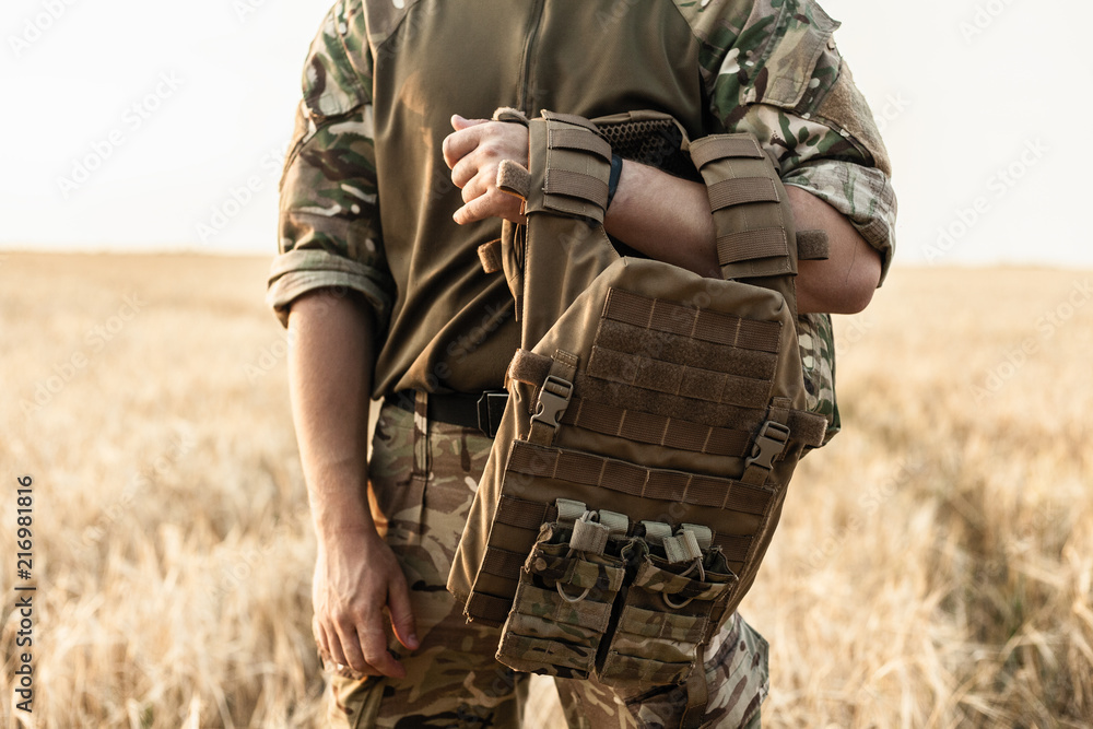 Soldier man standing against a field. Soldier in military outfit with bulletproof  vest. Photo of a soldier in military outfit holding a gun and bulletproof  vest on orange desert background. Stock Photo