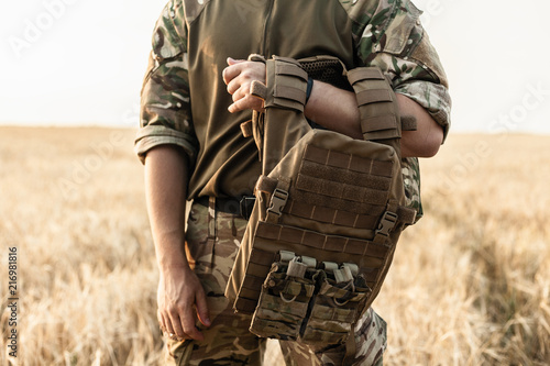 Soldier man standing against a field. Soldier in military outfit with bulletproof vest. Photo of a soldier in military outfit holding a gun and bulletproof vest on orange desert background. photo
