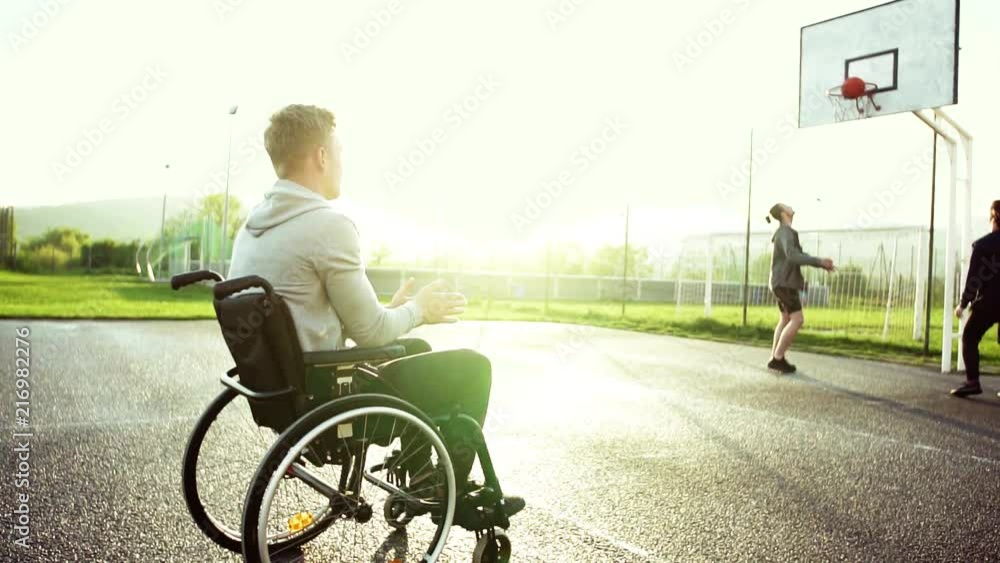 A teenager in wheelchair looking at boys playing basketball outdoors at sunset. A teenager in wheelchair looking at boys playing basketball outdoors at sunset.
