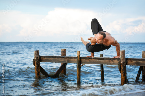 Young yoga trainer practicing yoga exercises on a wooden pier on a sea or river shore.