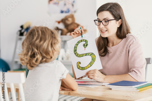 A child with development problems with a professional speech therapist during a meeting. Tutor holding a prop poster of a snake as a letter 's'.