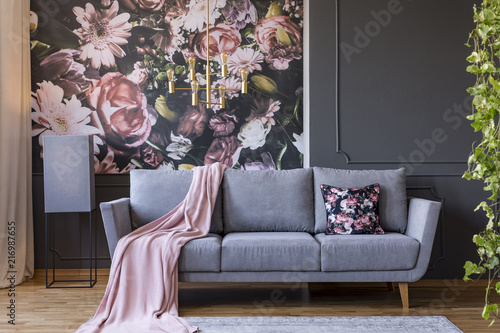 Powder pink blanket thrown on grey couch in real photo of dark sitting room interior with floral wallpaper, gold lamp and wainscoting on wall