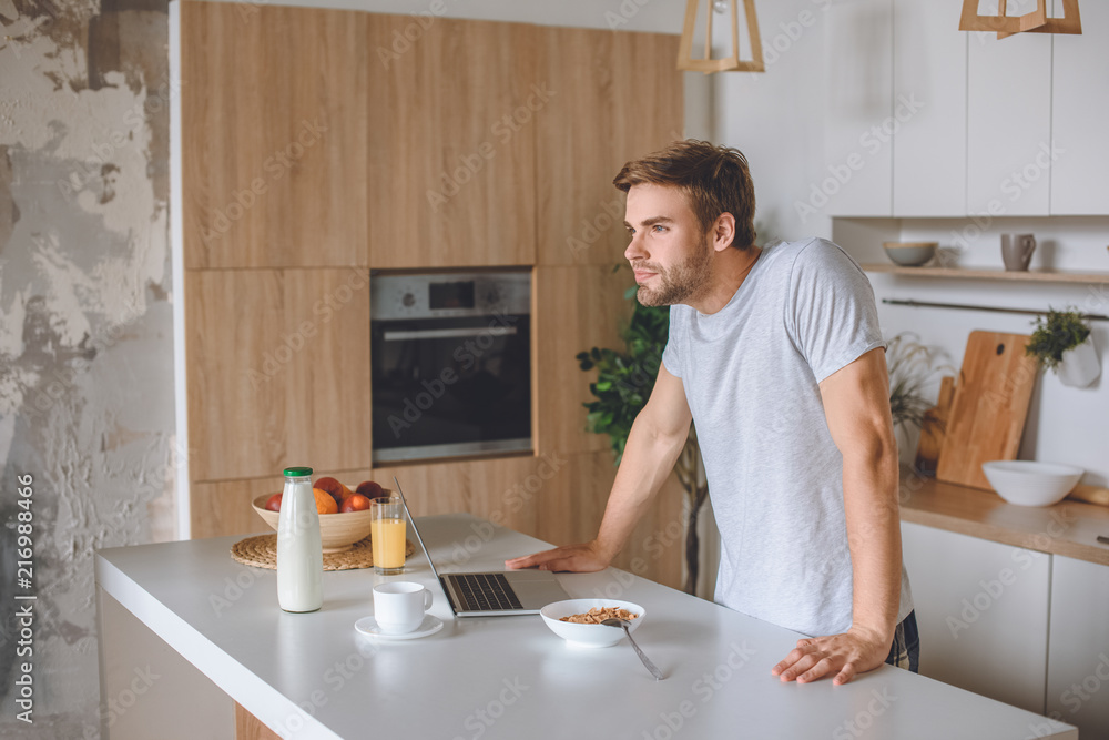 happy young man standing at table with breakfast and laptop at kitchen happy young man standing at table with breakfast and laptop at kitchen