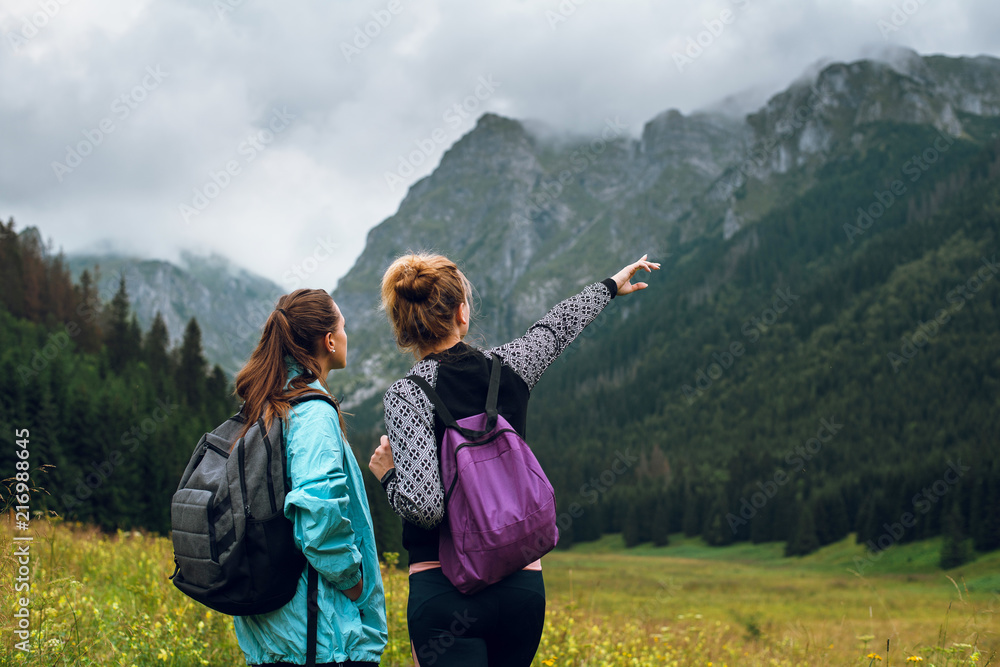 Rear view shot of two young women with backpacks enjoying beautiful view of green hills and rocky peaks in Rysy mountains, Tatras, Slovakia and Poland