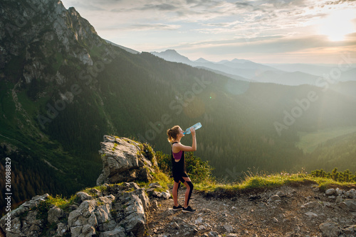 Young fit woman in sportswear looking drinking water on the mountain peak with scenic view of mountains on sunset. Rysy mountains, Tatras. Poland, Slovakia