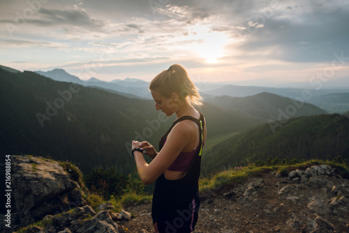 Side view of young sporty woman in sportswear using smartwatch while jogging in the mountains on sunset. High Tatra mountains